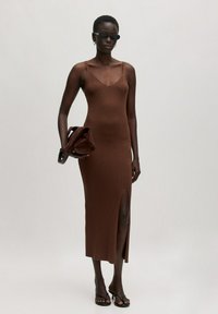 Bruine ribgebreide midi-jurk met dunne schouderbandjes en een hoge split tot boven de dij. Model houdt een bruine suede tas vast, draagt zwarte zonnebril en hakken.