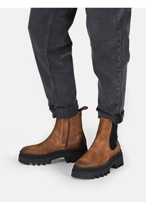 Brown suede ankle boots with a chunky black rubber sole, featuring a side zipper and elastic panels, worn with rolled black jeans.