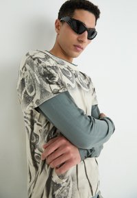 T-shirt graphique crème avec des motifs abstraits noirs, superposé à une chemise à manches longues teal. Des lunettes de soleil noires angulaires complètent le look.