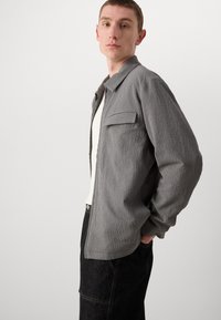 Chaqueta texturizada gris con cuello, un bolsillo en el pecho y cierre de botones, combinada con una camisa blanca y pantalones oscuros.