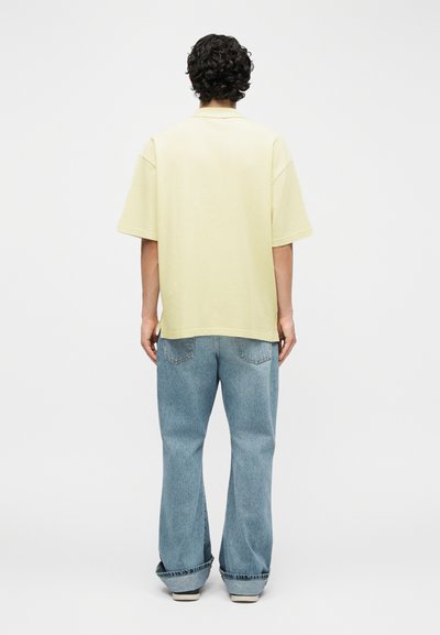 Άτομο με σγουρά σκούρα μαλλιά στέκεται με την πλάτη προς το μέρος μας, φορώντας ένα ανοιχτό κίτρινο κοντομάνικο πουκάμισο και φαρδιά μπλε τζιν με γυρισμένα μπατζάκια.