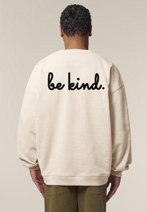 Sweatshirt beige oversize avec un col rond, ornée du texte "sois bienveillant" en cursive noir audacieux au dos. Texture de tissu douce.