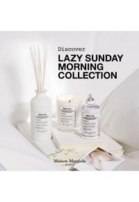 Tre Maison Margiela Replica parfymeprodukter på en hvit overflate med en bok og en blomst, som promoterer Lazy Sunday Morning-kolleksjonen.