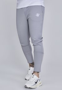 Lys grå atletikbukser med tætsiddende design, der har et logo på venstre hofte og kontraststing. Parret med hvide Nike sneakers.