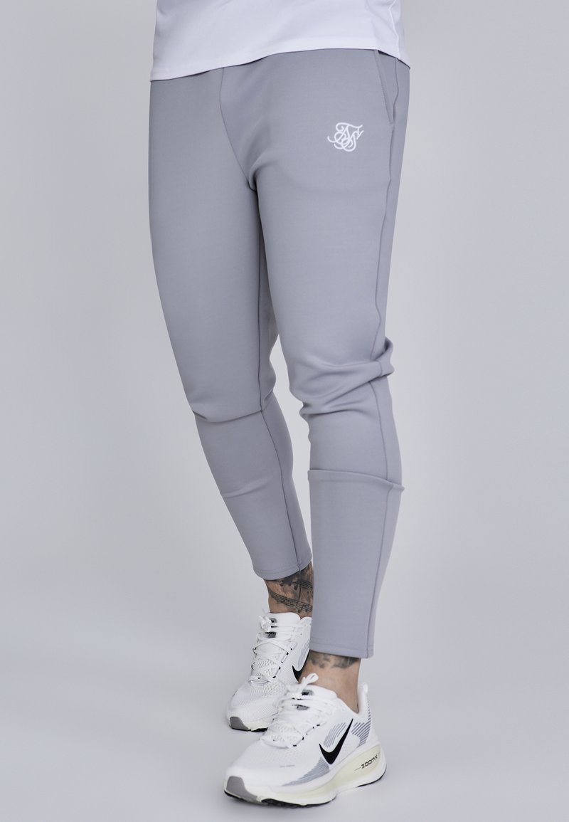 Lys grå atletikbukser med tætsiddende design, der har et logo på venstre hofte og kontraststing. Parret med hvide Nike sneakers.