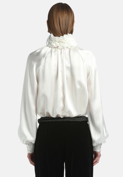Camicia di raso bianca con colletto arricciato e schiena elasticizzata. Maniche lunghe e voluminose con polsini abbottonati. Abbinata a pantaloni in velluto nero.