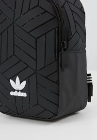 Svart Adidas-ryggsäck med geometriskt texturerat mönster, glansiga detaljer, dragkedjestängning och vit logotyp på framsidan. Justerbara remmar.