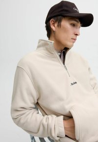 Young person wearing a beige fleece pullover with half-zip, black baseball cap, small hoop earring, seated with hands in front pocket.