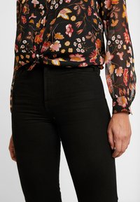 Floral black blouse with long, sheer sleeves tied at the waist, paired with high-waisted black jeans. Fabric features colorful floral patterns.