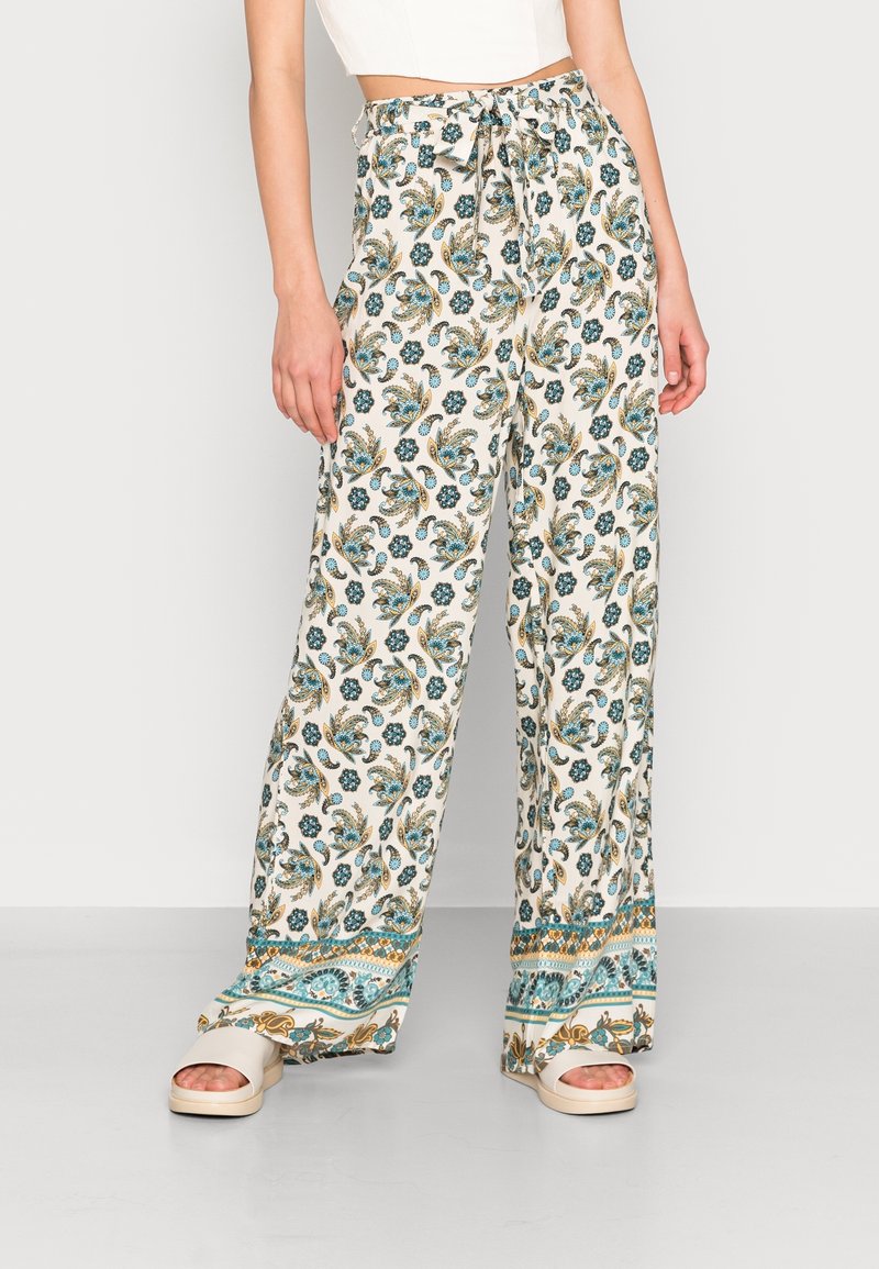 Femme portant un pantalon blanc à jambes larges avec un imprimé floral bleu et or et un ourlet à motif, associé à des sandales blanches à bouts ouverts.