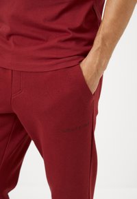 Burgundi színű melegítőnadrág, puha tapintással, oldalsó zsebbel és diszkrét fekete logónyomattal a lábszáron. Karcsú fazon.