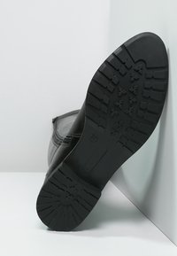 Svart läderstövel som visar en texturerad gummisula med djupa spår, som framhäver hållbarhet och greppfunktioner. Slät ovandel med dragkedjedetalj.