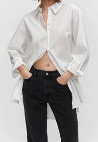 Camisa blanca de botones con un ajuste relajado, con mangas enrolladas y un diseño ligeramente recortado. Combinada con pantalones vaqueros negros de cintura alta.