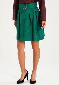 Gonna plissettata verde con tessuto testurizzato, cintura larga con fibbia circolare, abbinata a tacchi alti neri.