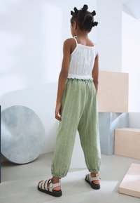 Light green striped pants with elastic cuffs and a white frilled tank top, paired with beige strappy sandals. Back view, minimal background.