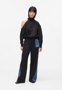 Sudadera negra con corte asimétrico y cuello alto, combinada con pantalones de pierna ancha que tienen un parche de denim en un lado.