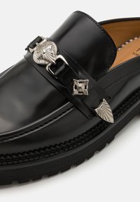 Mocasini din piele neagră, dotate cu o cataramă decorativă din argint și accente, cu o talpă din cauciuc texturat și cusături detaliate de-a lungul marginii.