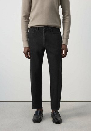 Hombre con vaqueros negros de corte recto, suéter beige de manga larga y mocasines de cuero negro, de pie frente a un fondo liso.