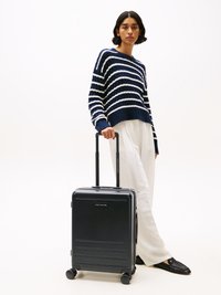Black hard-shell suitcase with embossed lines, retractable handle, and four wheels. Model wearing a navy and white striped sweater and light pants.