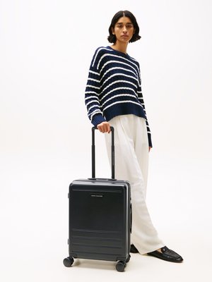 Black hard-shell suitcase with embossed lines, retractable handle, and four wheels. Model wearing a navy and white striped sweater and light pants.