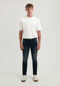 Bílá bavlněná tričko, kulatý výstřih, krátké rukávy; tmavě modré džíny, slim fit, vybledlý design; bílé tenisky s hnědými podrážkami. Neutrální pozadí.