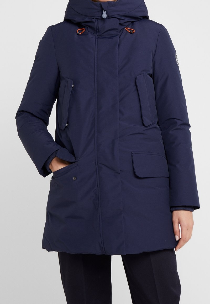 Manteau d'hiver isolé bleu marine avec capuche, accents de cordon orange, plusieurs poches à rabat et un écusson logo sur le côté, porté par une personne.