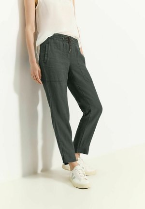 Personne portant un pantalon noir à cordon avec des poches latérales et des baskets blanches, debout contre un mur blanc les mains dans les poches.