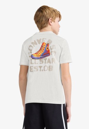 Blond chłopiec w białej koszulce, kolorowym wysokim trampku i wyblakłym napisie Converse All Star na plecach, w zestawieniu z czarnymi szortami.