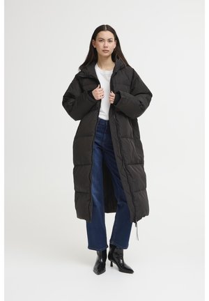 Zwarte lange pufferjas met rits aan de voorkant, gestikt ontwerp en een capuchon. Gecombineerd met een wit shirt en blauwe jeans, in combinatie met zwarte haklaarzen.