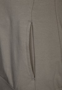 Šedá látka s jemnou texturou obsahující boční kapsu s prošitým švem. Otvor kapsy je strategicky umístěn na oděvu.