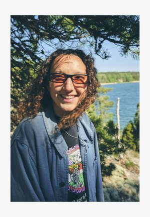 Barna göndör hajú személy sötét szemüveget visel és egy laza kék dzsekit fekete grafikus póló fölött, mosolyogva egy vízparti szabadtéri helyszínen.