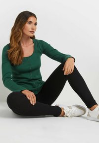 Vihreä ribbitoppi, jossa on pitkät hihat, neliökaula-aukko ja nappiyksityiskohtia, yhdistettynä mustiin skinny-housuihin ja valkoisiin tennareihin.