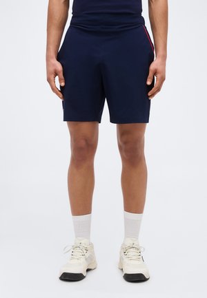 Homme portant un short de sport bleu marine, des chaussettes blanches montantes et des baskets blanches, debout devant un fond clair uni.