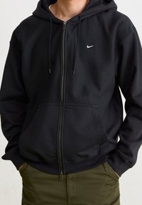 Svart zip-up hoodie med en framficka och dragsko i huvan. Har en liten vit Nike-logga på vänster bröst. Mjuk tyg, avslappnad passform.