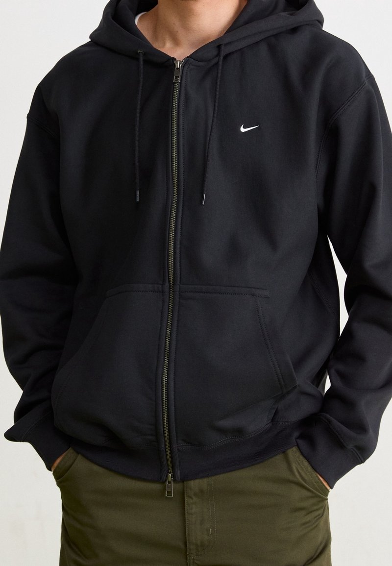 Svart zip-up hoodie med en framficka och dragsko i huvan. Har en liten vit Nike-logga på vänster bröst. Mjuk tyg, avslappnad passform.