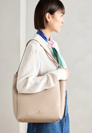 Femme en haut blanc et foulard coloré portant un sac à bandoulière beige avec des détails dorés, debout devant un fond clair uni.