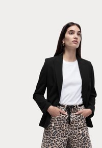 Černý tailored blazer přes bílý tričko, spárovaný s béžovými kalhotami s leopardím potiskem. Model má dlouhé tmavé vlasy a nosí zlaté kruhové náušnice.