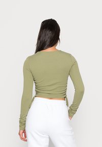 Vihreä pitkähihainen paita, jossa on istuva muotoilu, takana gathered-yksityiskohtia ja nauhat sivuilla. Yhdistetty valkoisiin housuihin.