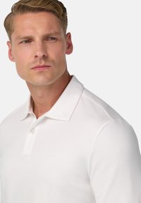 Balta garām piedurknēm polo krekls ar apkakles izgriezumu, vienu pogu un gludu tekstūru. Tuvplāns skatījums uz augšējo ķermeņa daļu.