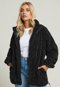 Musta, tekstuuri fleece-takki hupulla, ylisuuri istuvuus ja näkyvät taskut. Varustettu vetoketjukiinnityksellä ja huomaamattomalla timanttikuvioinnilla.