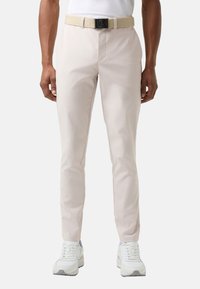 Jasnobeżowe spodnie chino o wąskim kroju, wykonane z teksturowanej tkaniny, z szlufkami na pasek i dołączonym paskiem w tym samym kolorze. Noszone z białymi trampkami.