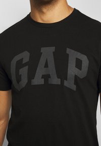 Camiseta de algodón negra con un gran logo "GAP" en gris oscuro en el frontal. El diseño tiene cuello redondo y mangas cortas.