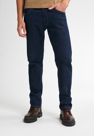 Jeans de mezclilla azul oscuro con un diseño de pierna recta, que cuentan con un bolsillo frontal y costuras visibles. Combinados con zapatos marrones.