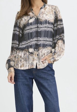 Kvinde iført en langærmet sort og beige paisley-mønstret bluse, der er stukket ud over blå jeans, med den ene hånd i lommen.