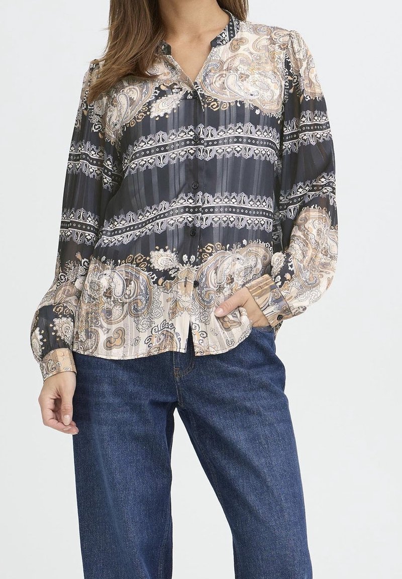 Femme portant un chemisier à manches longues noir et beige à motif cachemire, porté sur un jean bleu, avec une main dans la poche.