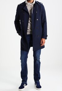 Trench coat doppiopetto blu navy con spalline sulle spalle, abbinato a una felpa con cappuccio grigia, jeans blu e sneakers blu navy con dettagli bianchi.