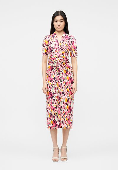 Femme portant une robe mi-longue à manches courtes, avec un froncé au centre et un motif abstrait multicolore rose, jaune et noir, debout devant un fond blanc.