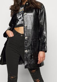 Abrigo de charol negro con textura de cocodrilo, que presenta botones plateados y bolsillos, combinado con jeans negros rasgados y una camiseta gráfica corta.