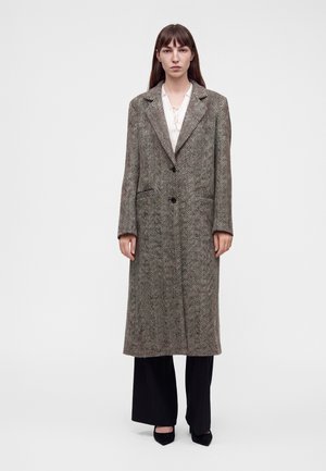 Mujer de pie con un abrigo largo marrón de espiga encima de una blusa blanca, pantalones anchos negros y zapatos puntiagudos negros sobre un fondo blanco.