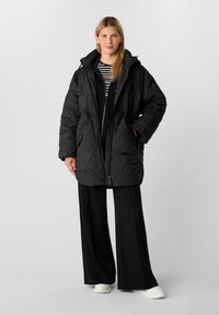 Schwarze gesteppte Jacke mit Kapuze, ausgestattet mit einem Frontreißverschluss, einer verstellbaren Taille und großen Taschen. Getragen über schwarzen Hosen und einem gestreiften Oberteil.